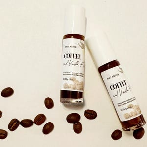 May include: Two brown glass roller bottles with white labels. The labels say "ANTI AGING COFFEE and Vanilla Face" and "MADE WITH ORGANIC COFFEE, SAFFLOWER, MEADOWFOAM OIL 28.35 g/1 oz". The bottles are surrounded by coffee beans on a white background.