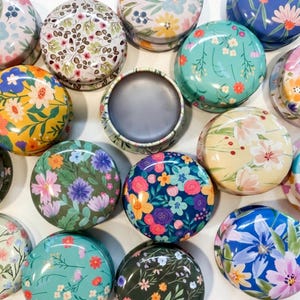 May include: A collection of round metal tins featuring various colorful floral patterns, including yellow, teal, dark blue, and cream backgrounds with printed flowers in shades of pink, orange, purple, and green, with one tin shown open.
