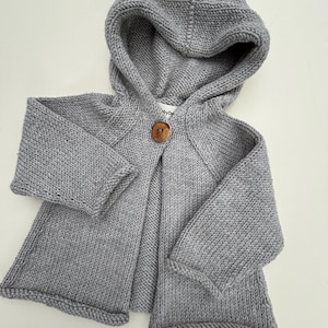 May include: A light grey, hooded knitted cardigan with a single wooden button fastening. The cardigan has long sleeves and a cosy, textured knit design. The garment is designed for children.