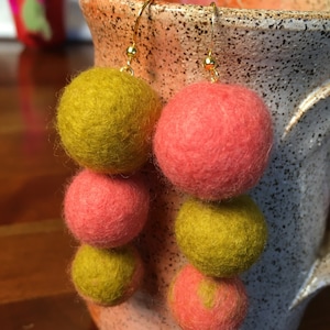 Puede incluir: Un par de pendientes colgantes con tres bolas de fieltro en cada pendiente. La bola superior es amarilla, la bola del medio es rosa y la bola inferior es amarilla. Los pendientes están colgados de una taza.