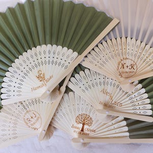 May include: A collection of personalized wooden fans with intricate cut-out designs. The fans feature names and dates, with a green and white background. The text includes names like Hannah Blake, Alex Lara, and Isaac Olivia, along with dates.