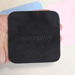 Puede incluir: Un pa&ntilde;o de microfibra cuadrado negro con el texto "SusieCreative" en relieve en blanco.