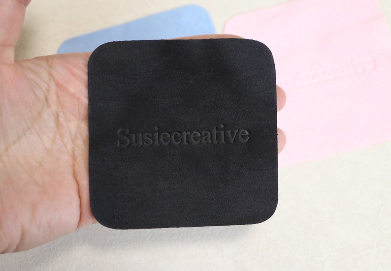 Puede incluir: Un pa&ntilde;o de microfibra cuadrado negro con el texto "SusieCreative" en relieve en blanco.