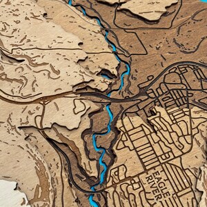 May include: A detailed wooden map with laser-cut layers, showcasing a topographical view. The map features a river highlighted in turquoise, with the text "EAGLE RIVER" visible. The wood has varying shades of brown, creating a 3D effect.
