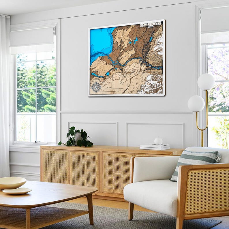 May include: A framed wooden map of Eagle River, with a detailed relief of the terrain. The map features shades of blue for water and brown for land, with the text "EAGLE RIVER" at the top. The artwork is displayed in a bright, modern living room.