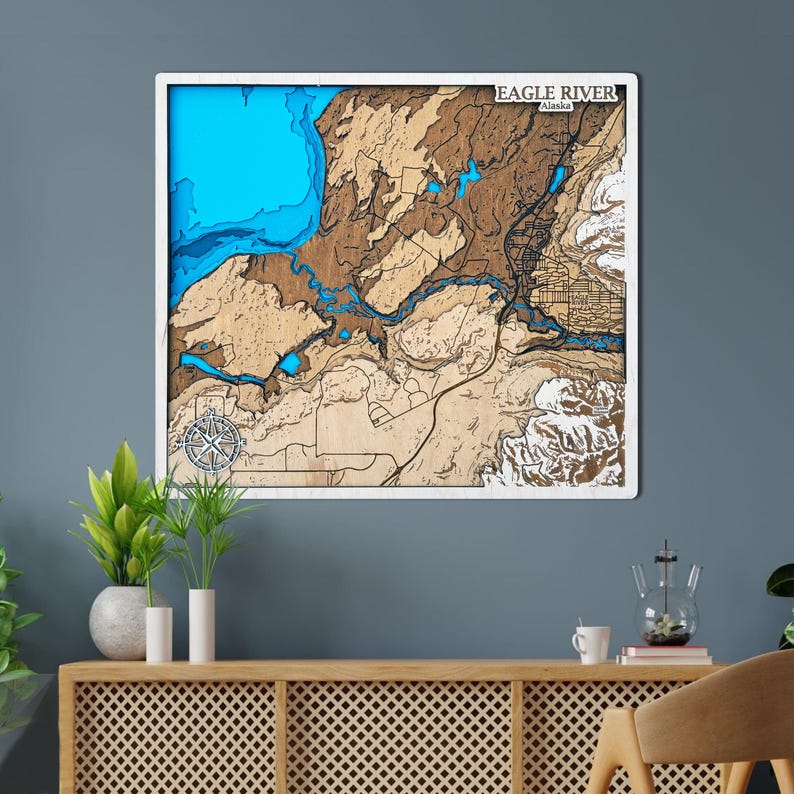 May include: A wooden topographic map of Eagle River, Alaska, framed in white. The map features layered details in shades of brown, blue, and white, with a compass rose and the text "EAGLE RIVER Alaska". The artwork is displayed on a blue wall.