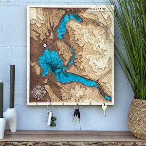 May include: Wooden wall-mounted key holder featuring a detailed topographic map with a blue lake. The map is framed in a light wood border and includes a compass design. It has several key hooks and the text "ARAPAHO National Recreation Area".