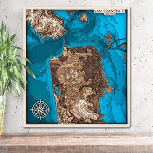 May include: A framed, multi-layered wooden map of San Francisco, California, with a detailed depiction of the city's topography and surrounding waters. The map features a compass rose and the text "SAN FRANCISCO California".