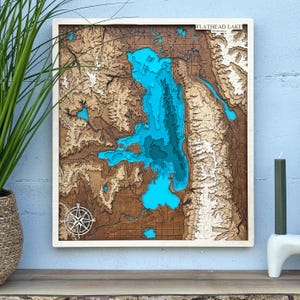 May include: A wooden topographic map of Flathead Lake, Montana, with a white frame. The lake is depicted in shades of blue, surrounded by detailed brown and tan terrain. A compass rose is in the lower left corner. The text "FLATHEAD LAKE" is at the top.