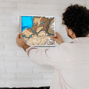 May include: A framed wooden map of Eagle River, with a white border. The map features a blue lake, brown land, and the text "EAGLE RIVER" at the top. The map is being held against a white brick wall.