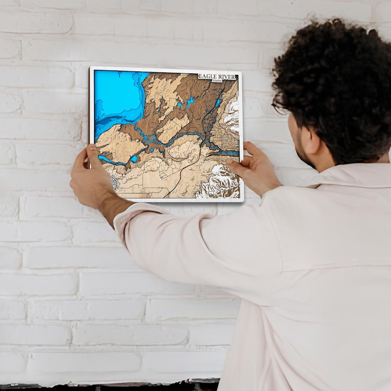 May include: A framed wooden map of Eagle River, with a white border. The map features a blue lake, brown land, and the text "EAGLE RIVER" at the top. The map is being held against a white brick wall.