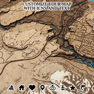 May include: A layered wooden map with the text "CUSTOMIZE YOUR MAP WITH ICNS AND TEXT". The map features a detailed depiction of a landscape with rivers and the text "EAGLE RIVER". The map has a natural wood grain finish with blue accents.