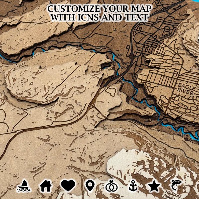 May include: A layered wooden map with the text "CUSTOMIZE YOUR MAP WITH ICNS AND TEXT". The map features a detailed depiction of a landscape with rivers and the text "EAGLE RIVER". The map has a natural wood grain finish with blue accents.