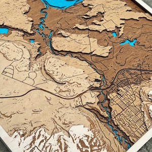 May include: A wooden topographic map with laser-cut layers. The map features a detailed depiction of a geographical area, with rivers and lakes highlighted in blue. The map is framed in a light-colored wood.