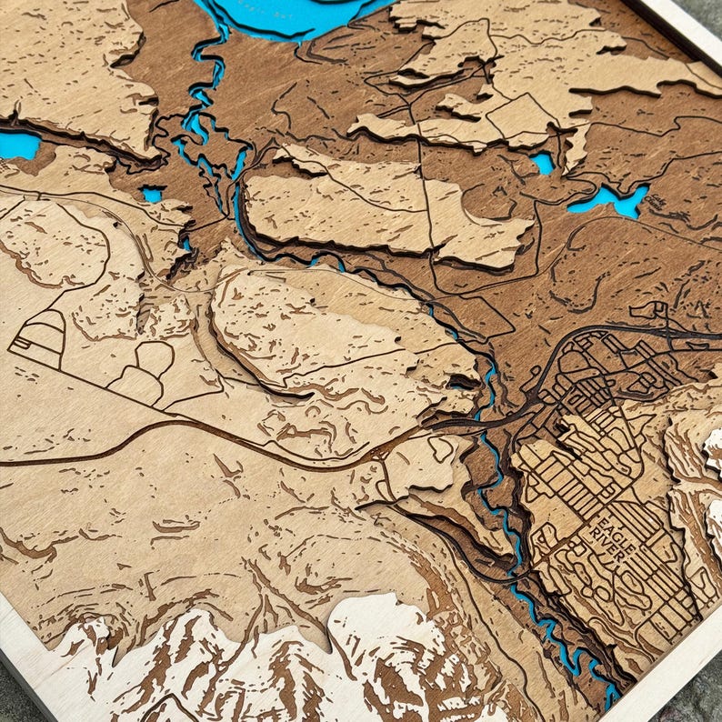 May include: A wooden topographic map with laser-cut layers. The map features a detailed depiction of a geographical area, with rivers and lakes highlighted in blue. The map is framed in a light-colored wood.