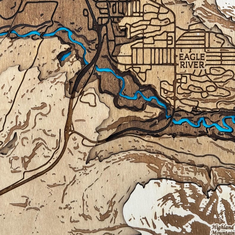 May include: A layered wooden map with a detailed topographical design. The map features a river in a vibrant blue color, with the text "EAGLE RIVER" visible. The map is made of various shades of brown wood.