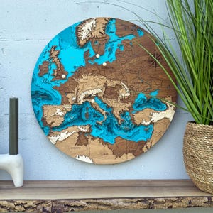 Könnte beinhalten: Eine runde Holzkarte von Europa mit detailliertem, geschichtetem Design. Die Karte zeigt Brauntöne für das Land und Türkis für das Wasser, mit in das Holz geätzten Ländernamen. Eine Windrose ist sichtbar.