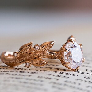 May include: A rose gold engagement ring featuring a large, round, clear diamond set in a delicate, claw-like setting. The band is adorned with a detailed, leafy design with smaller diamonds set into the leaves.