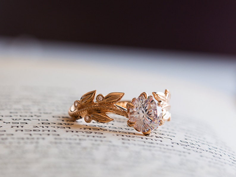 May include: A gold ring featuring a large, clear diamond and a delicate leaf design. The ring is resting on a page of a book.