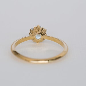 Puede incluir: Un anillo de oro con una piedra preciosa azul claro. El anillo presenta un dise&ntilde;o floral alrededor de la piedra preciosa y una banda detallada. El anillo est&aacute; sobre un fondo blanco, resaltando su elegante dise&ntilde;o.