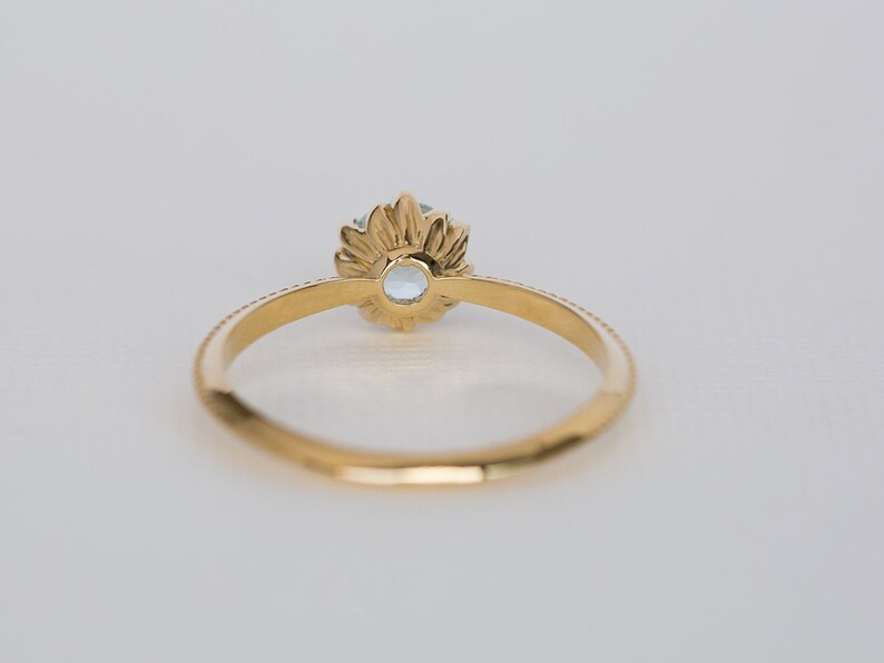 Puede incluir: Un anillo de oro con una piedra preciosa azul claro. El anillo presenta un dise&ntilde;o floral alrededor de la piedra preciosa y una banda detallada. El anillo est&aacute; sobre un fondo blanco, resaltando su elegante dise&ntilde;o.