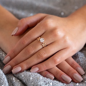 May include: A gold ring with a large, round, clear gemstone set in a delicate, leaf-shaped band. The ring is worn on the ring finger of a hand with light pink nail polish.