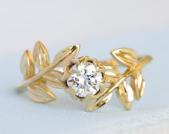May include: A gold ring featuring a central round brilliant-cut clear gemstone held in a floral-inspired setting, flanked by two symmetrical branches with polished gold leaves extending along the band on both sides of the center stone.