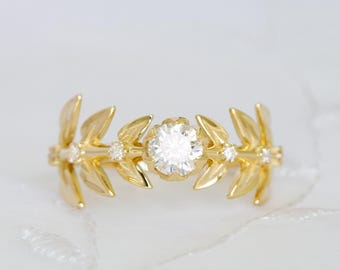 May include: A gold ring featuring a central round brilliant-cut white gemstone flanked by symmetrical leaf-shaped metal motifs. The band incorporates four smaller round white accent stones embedded within the golden foliage design.