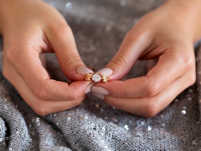 May include: A close-up of a hand wearing a gold ring with a large, clear diamond. The ring is set in a delicate, floral design.
