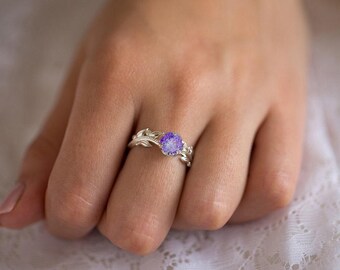 May include: A silver ring featuring a round, faceted purple stone set in a prong setting. The band is designed with a detailed metallic leaf and vine motif that wraps around the finger, creating a nature-inspired look with a polished finish.