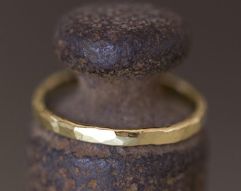 May include: A thin, gold-coloured ring with a hammered, textured surface. The band features a reflective, metallic finish with irregular facets along its circumference, creating a light-catching effect across the entire outer surface of the ring.