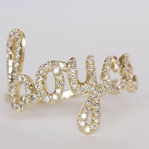 Puede incluir: Un anillo de oro con la palabra "boyce" escrita en una fuente cursiva. Toda la superficie de las letras está incrustada con pequeñas piedras claras y brillantes que captan la luz, creando un aspecto luminoso y reflectante sobre el metal.