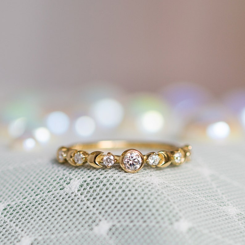 K&ouml;nnte beinhalten: Ein zarter Goldring mit einer Reihe kleiner Diamanten und einem gr&ouml;&szlig;eren Diamanten in der Mitte. Der Ring liegt auf einem hellgr&uuml;nen Stoffhintergrund.