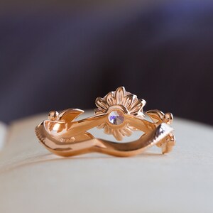 May include: A gold ring featuring a sunflower design with a clear gemstone at its centre. The ring is resting on a white surface.