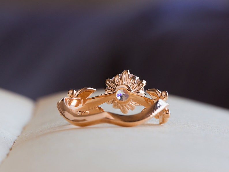 May include: A gold ring featuring a sunflower design with a clear gemstone at its centre. The ring is resting on a white surface.