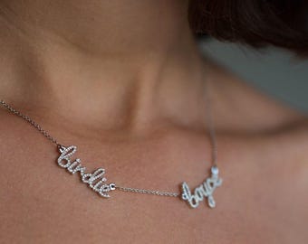 Op de afbeelding: Een zilverkleurige ketting met twee hangers in cursief schrift, "birdie" en "boyce", beide ingelegd met kleine doorzichtige steentjes. De fijne ketting verbindt de twee namen, wat zorgt voor een gelaagde look op het sleutelbeen.