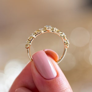 K&ouml;nnte beinhalten: Ein zarter Goldring mit einer Reihe kleiner, klarer Edelsteine. Der Ring wird von einer Hand mit rosafarbenem Nagellack gehalten.