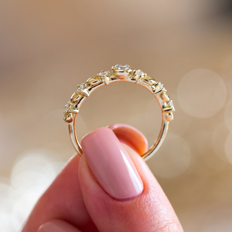 K&ouml;nnte beinhalten: Ein zarter Goldring mit einer Reihe kleiner, klarer Edelsteine. Der Ring wird von einer Hand mit rosafarbenem Nagellack gehalten.