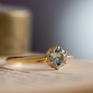 Puede incluir: Un anillo de oro con una gema azul claro. El anillo tiene un engaste detallado y una banda delgada. La gema redonda y facetada captura la luz. El anillo est&aacute; sobre una superficie de color claro.