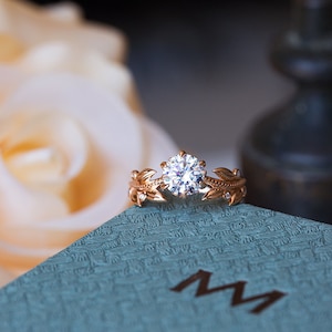 May include: A delicate gold ring featuring a large, round, clear diamond centre stone. The band is embellished with small diamonds and intricate leaf designs.