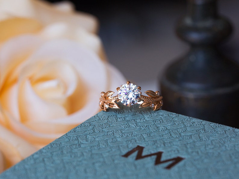 May include: A delicate gold ring featuring a large, round, clear diamond centre stone. The band is embellished with small diamonds and intricate leaf designs.