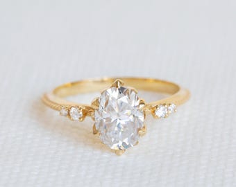 May include: A gold ring featuring a central oval-cut clear gemstone held in a prong setting. The band displays a textured rope-like design on the shoulders, accented by two small round clear stones on each side of the main oval setting.