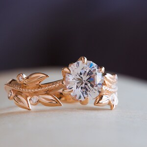 May include: A rose gold engagement ring featuring a large, round, clear diamond set in a floral design. The band is decorated with delicate leaves and smaller diamonds.