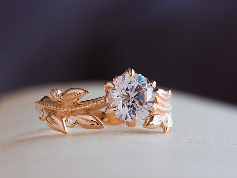 May include: A rose gold engagement ring featuring a large, round, clear diamond set in a floral design. The band is decorated with delicate leaves and smaller diamonds.
