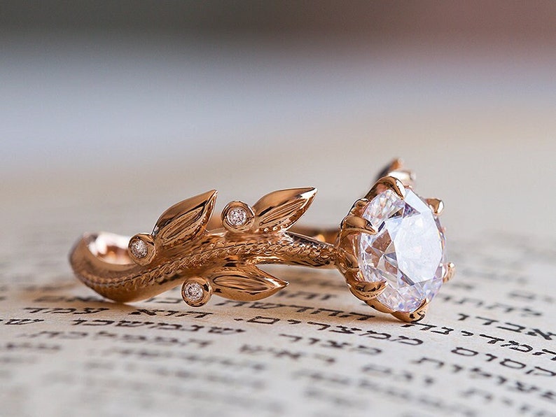 May include: A rose gold engagement ring featuring a large, round, clear diamond set in a delicate, claw-like setting. The band is adorned with a detailed, leafy design with smaller diamonds set into the leaves.