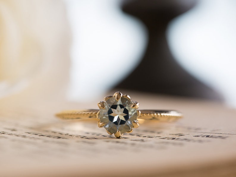 Puede incluir: Un anillo de oro con una piedra preciosa redonda y clara engastada en un engaste de garras floral. La banda tiene un dise&ntilde;o texturizado. El anillo est&aacute; sobre una p&aacute;gina de libro vintage.