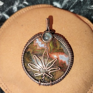 Könnte beinhalten: Ein handgefertigter Anhänger mit einem Kupferdrahtrahmen. Der Anhänger hat einen runden, mehrfarbigen Stein mit einem Cannabisblatt-Design aus Kupferdraht. Oben ist ein kleiner, runder, blau-grüner Stein angebracht.