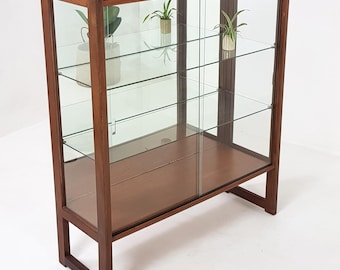 May include: A wooden display cabinet with glass doors and shelves. The cabinet has a rectangular shape with a dark brown finish. The glass doors and shelves are clear, allowing visibility of the contents. The cabinet is designed to showcase items.