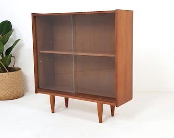 May include: A mid-century modern wooden display cabinet with glass sliding doors. The cabinet has two shelves and sits on four tapered legs. The wood has a warm, reddish-brown tone, and the glass doors are tinted.