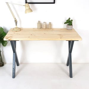 May include: A light-coloured wooden desk with dark grey crisscross legs. The desk has a rectangular top and is accessorised with a desk lamp, small bottles, and a potted plant. The desk is approximately 122 cm long.